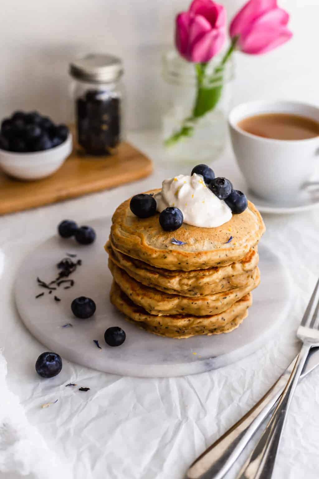 Stack of fluffy Earl Grey pancakes topped with whipped cream and blueberries, served with a cup of tea and fresh flowers in the background.