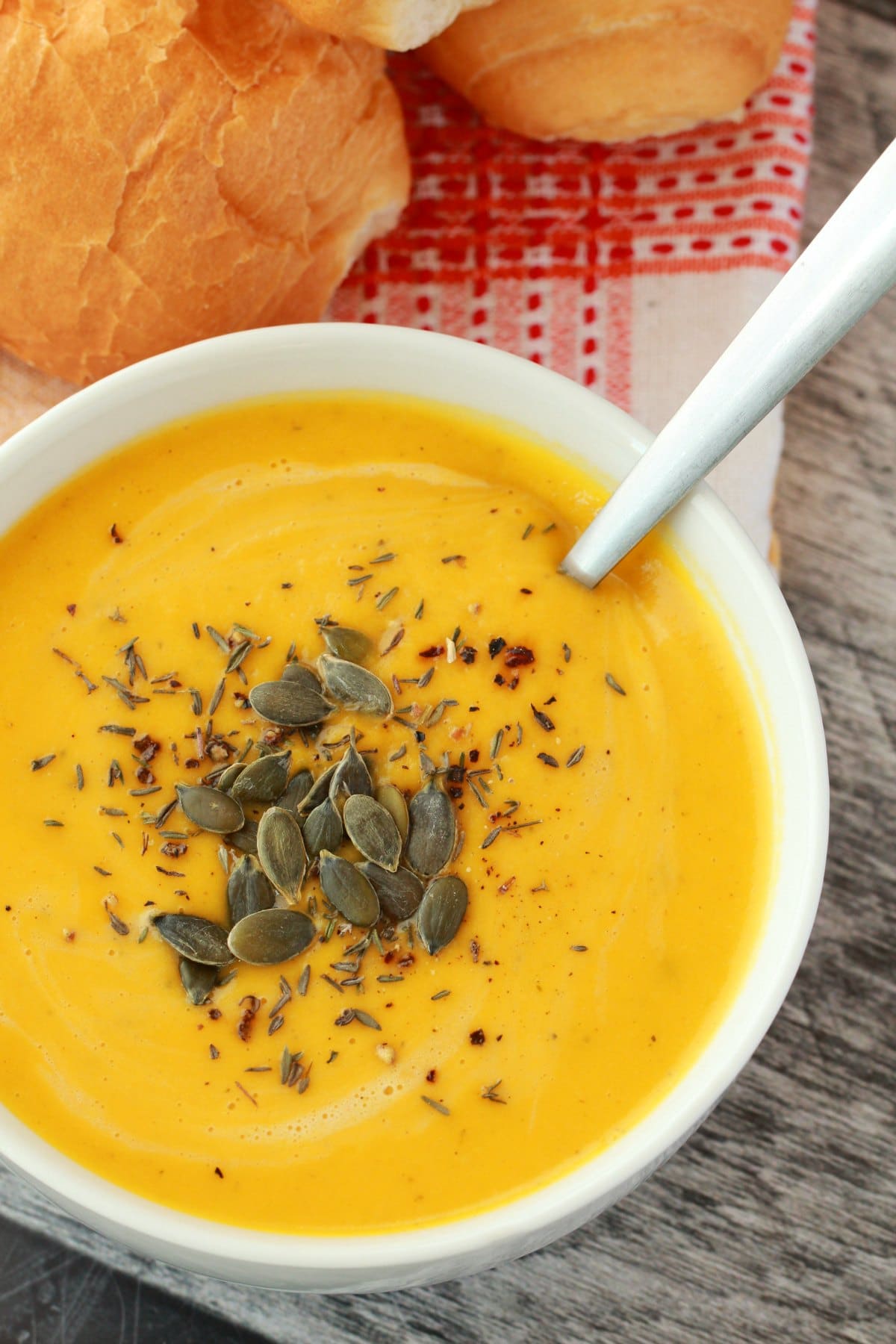 Bowl of creamy vegan pumpkin soup topped with pumpkin seeds and herbs, served with bread rolls on the side.