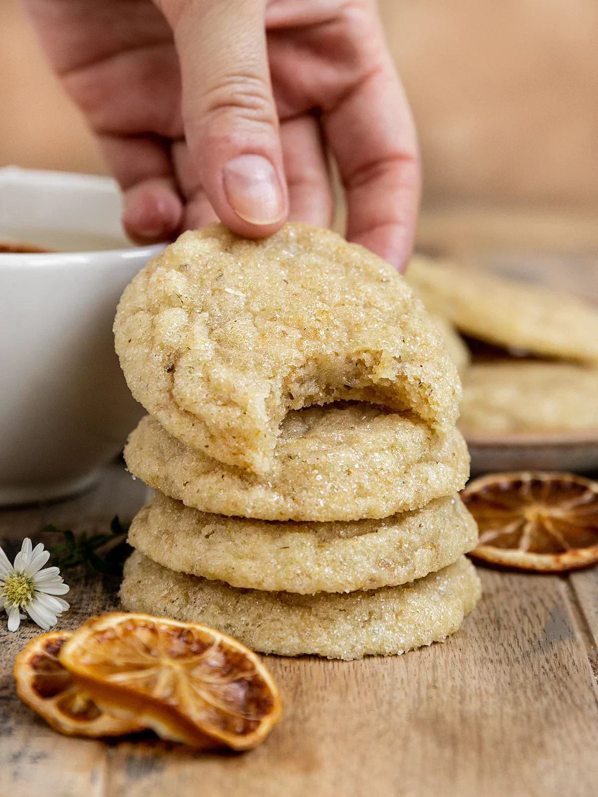 Hand holding a stack of soft vegan chamomile sugar cookies with one cookie showing a bite, next to dried citrus slices and a cup of tea.